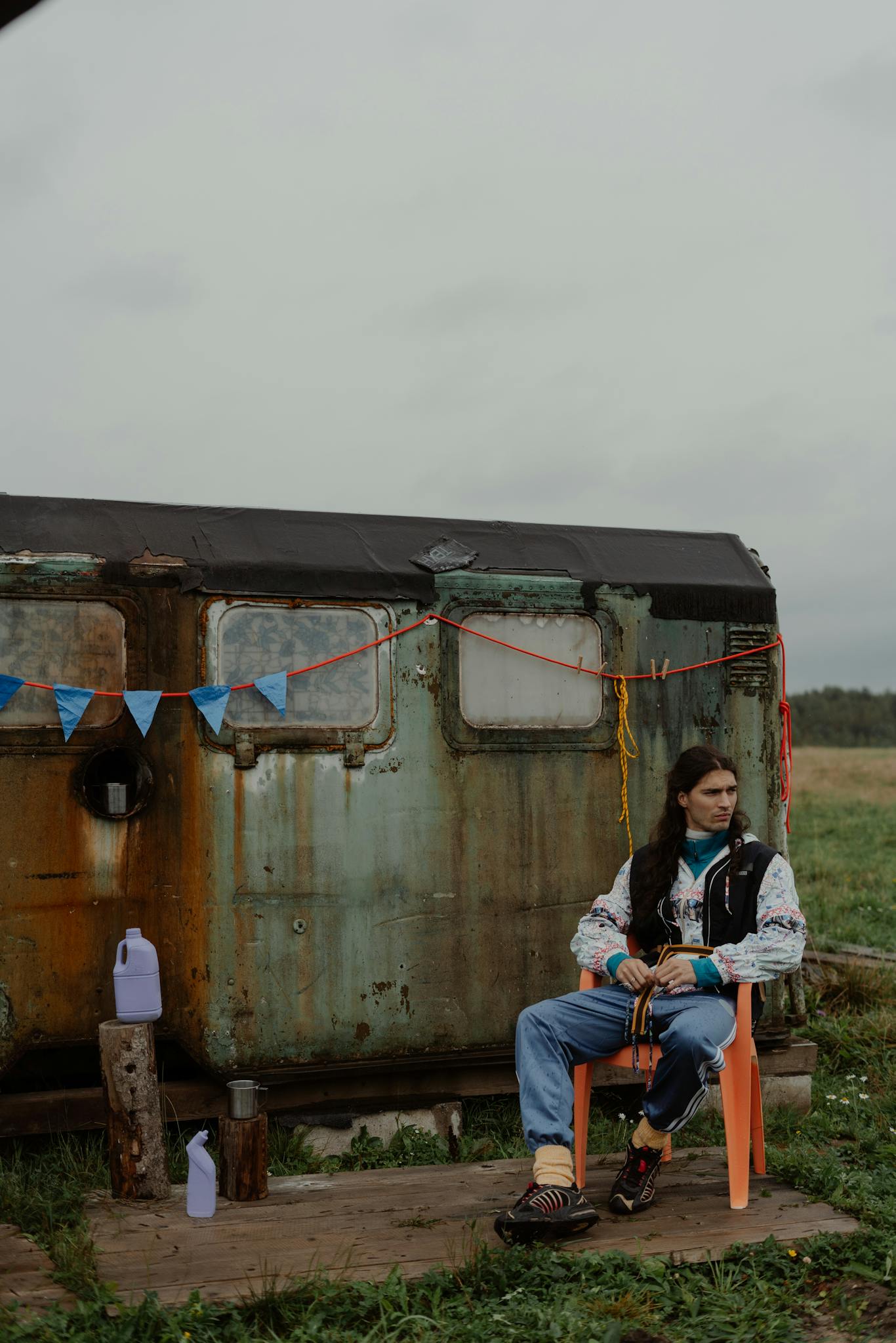 A young man embraces nomadic living, seated by a rustic camper in a grassy field.